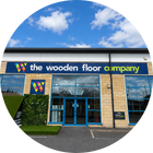The exterior of 'The Wooden Floor Company' building under a blue sky with white clouds, shown in a circular crop. The building features a blue sign with a colorful geometric logo, a beige brick facade, and large glass windows.